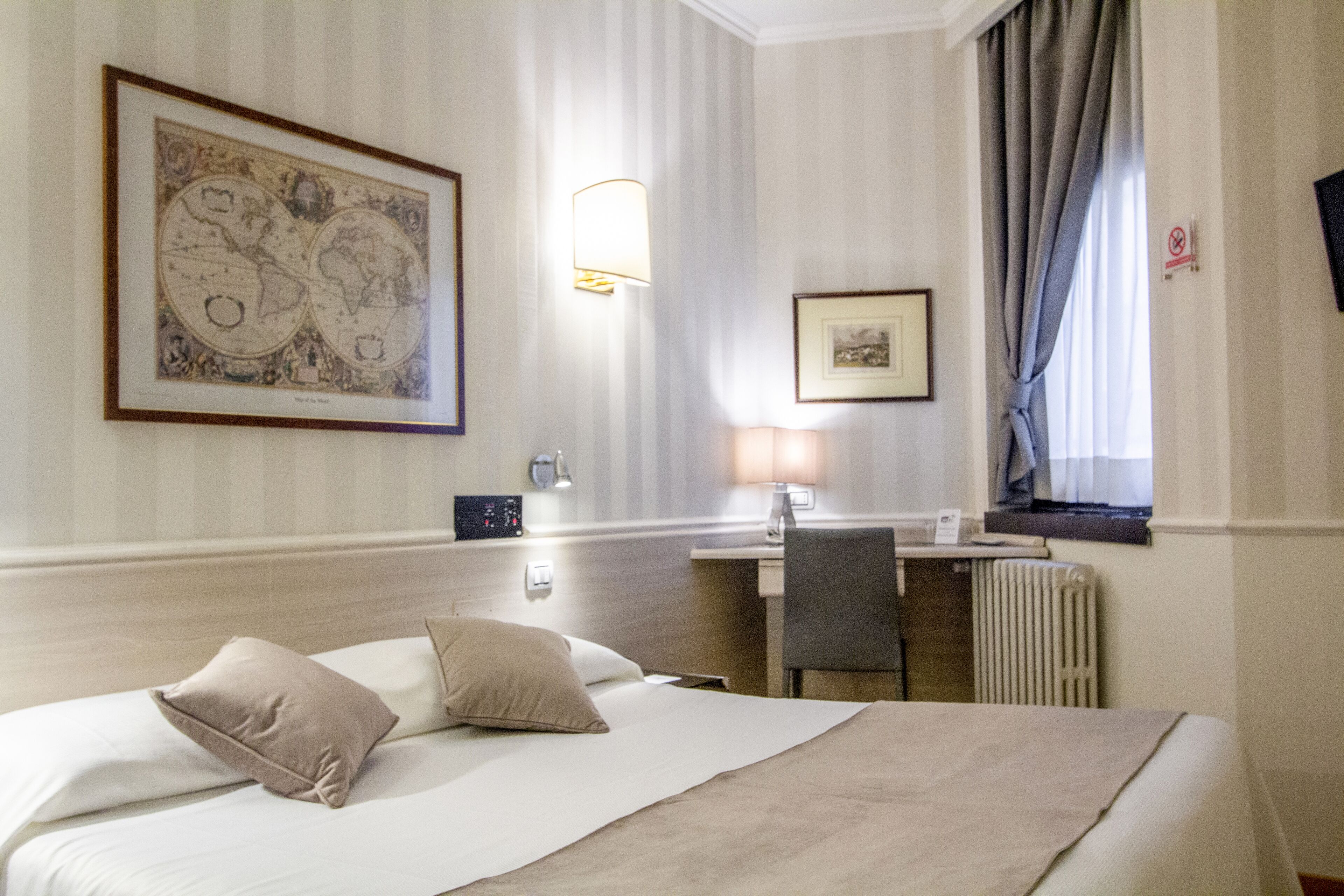 A hotel room with a bed, a desk, a chair, a lamp, a framed world map, and a window with curtains.