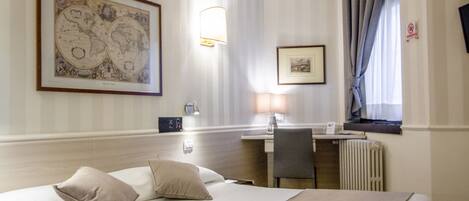 A hotel room with a bed, a desk, a chair, a lamp, a framed world map, and a window with curtains.