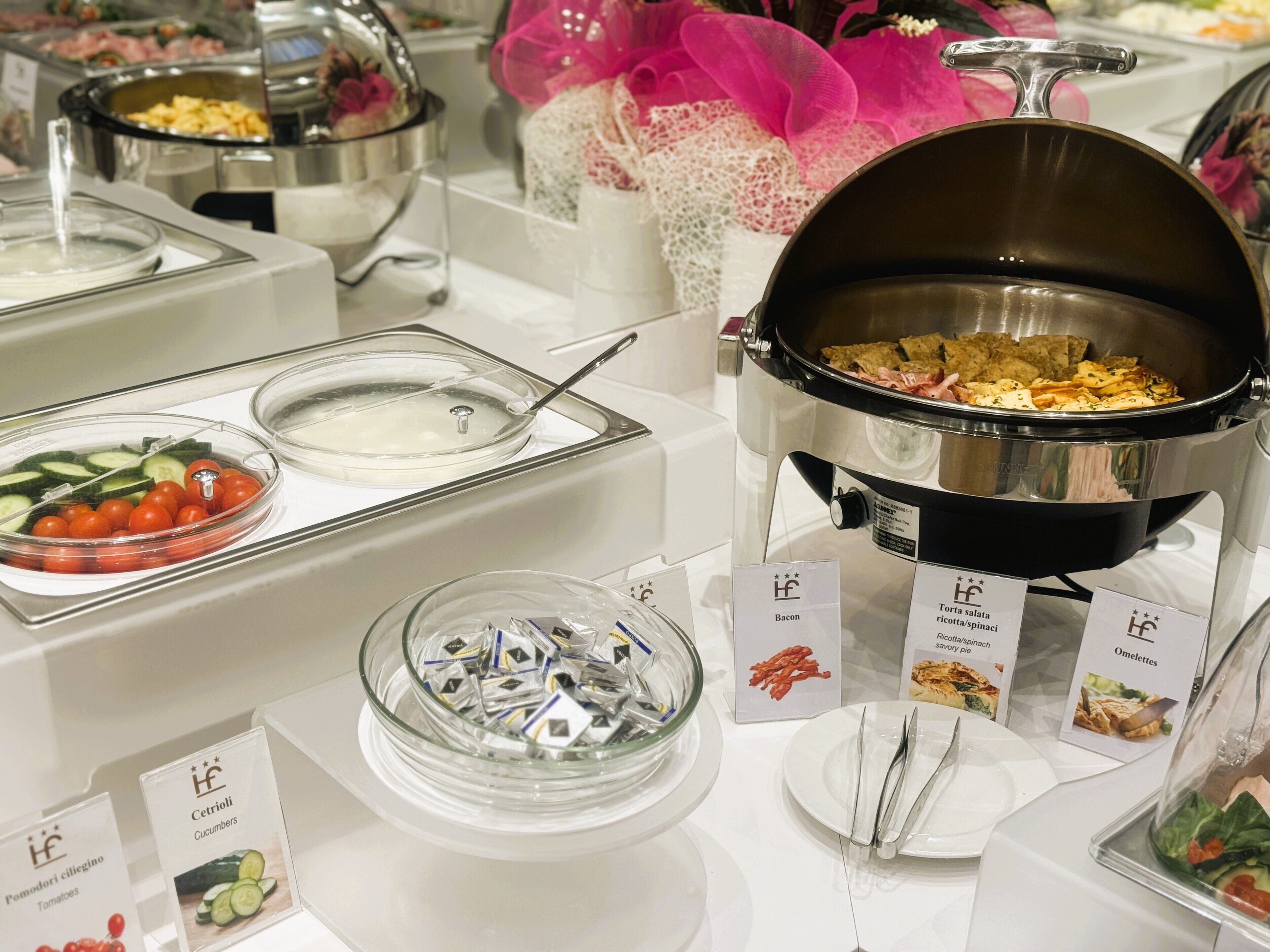 A buffet with various food options including tomatoes, cucumbers, bacon, omelettes, and a hot pot.
