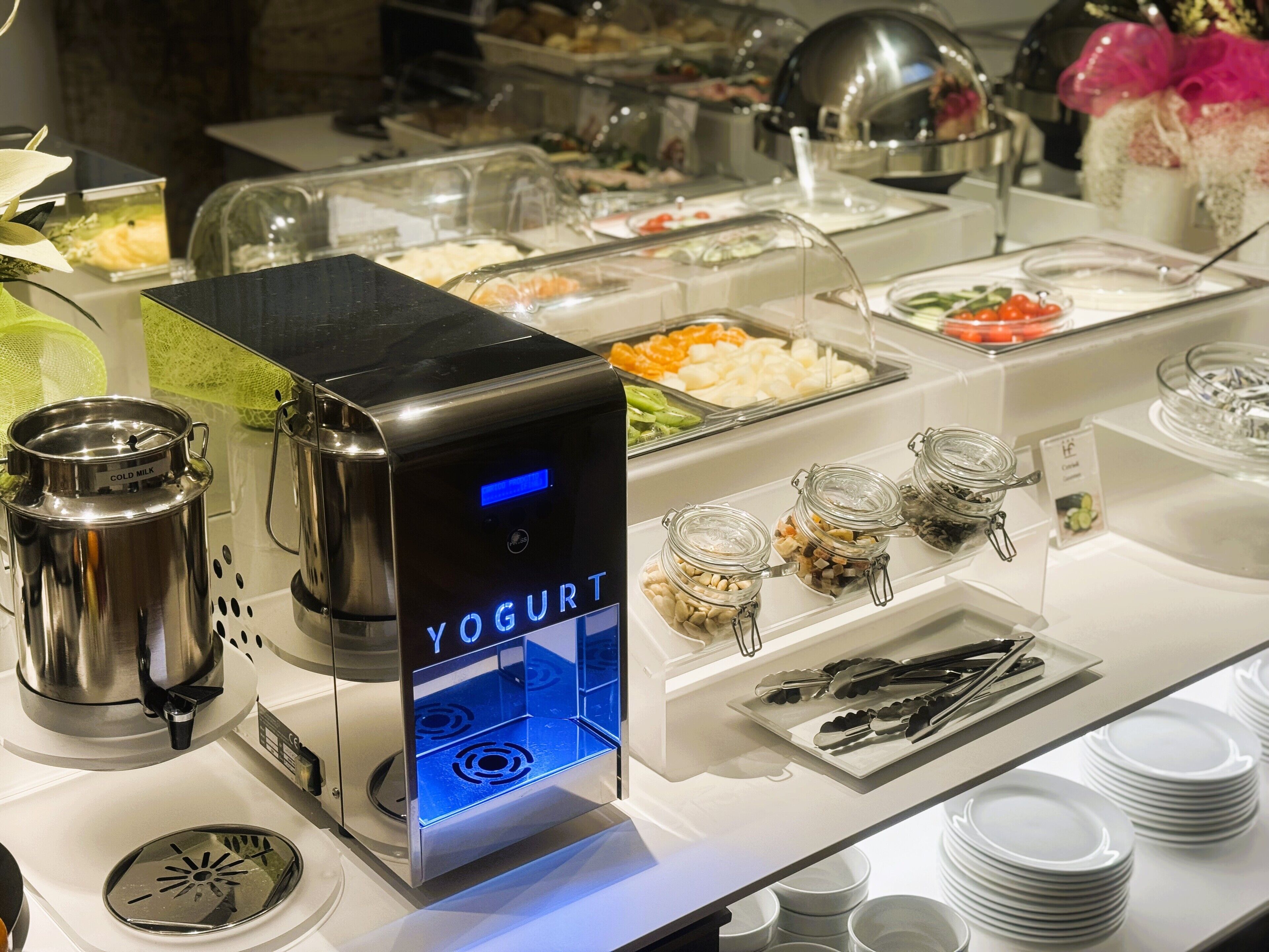 A buffet setup with a yogurt dispenser, various food containers, and utensils.