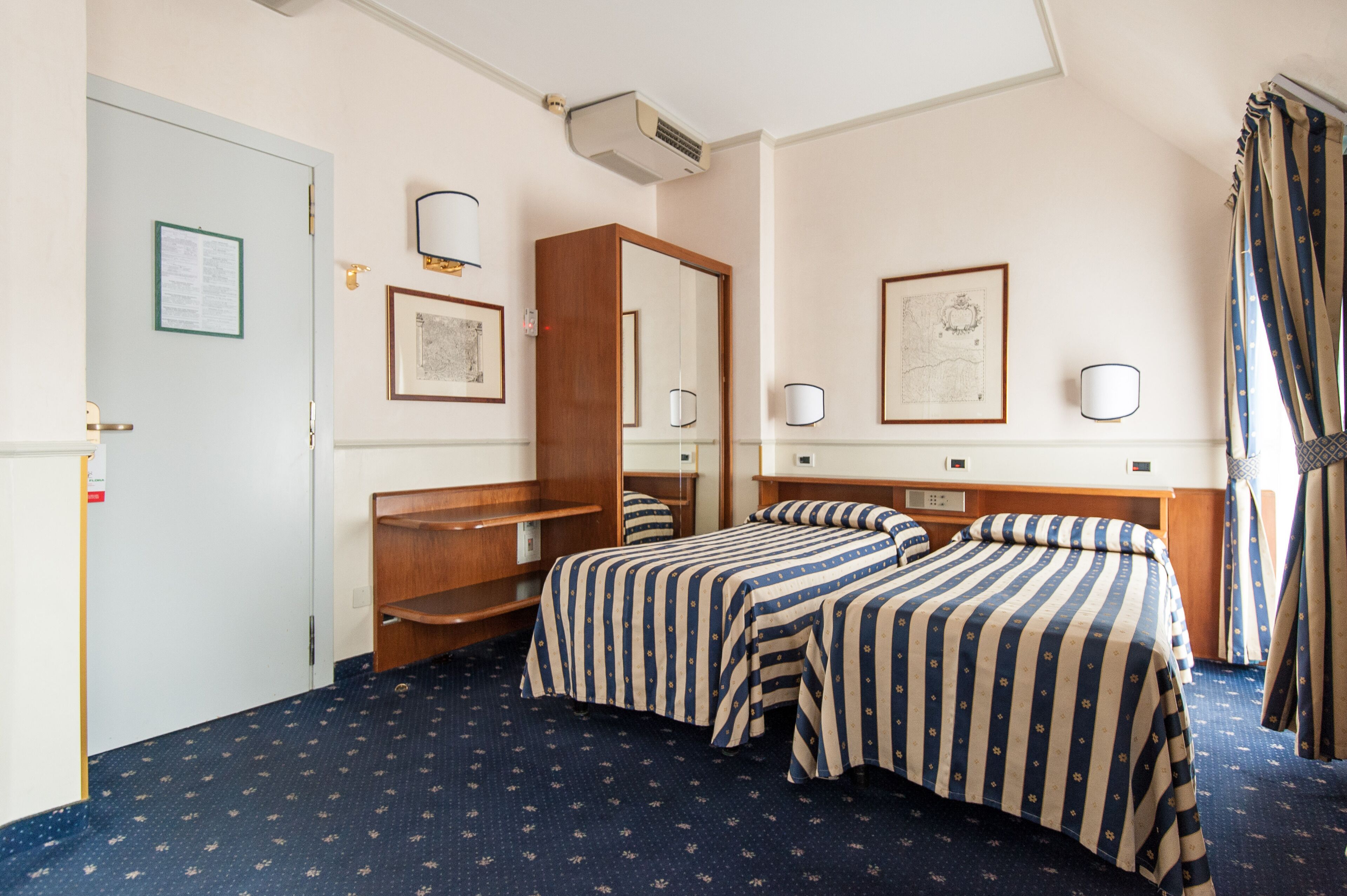 A hotel room with two beds, a wooden wardrobe, a mirror, framed artwork, and a window with curtains.