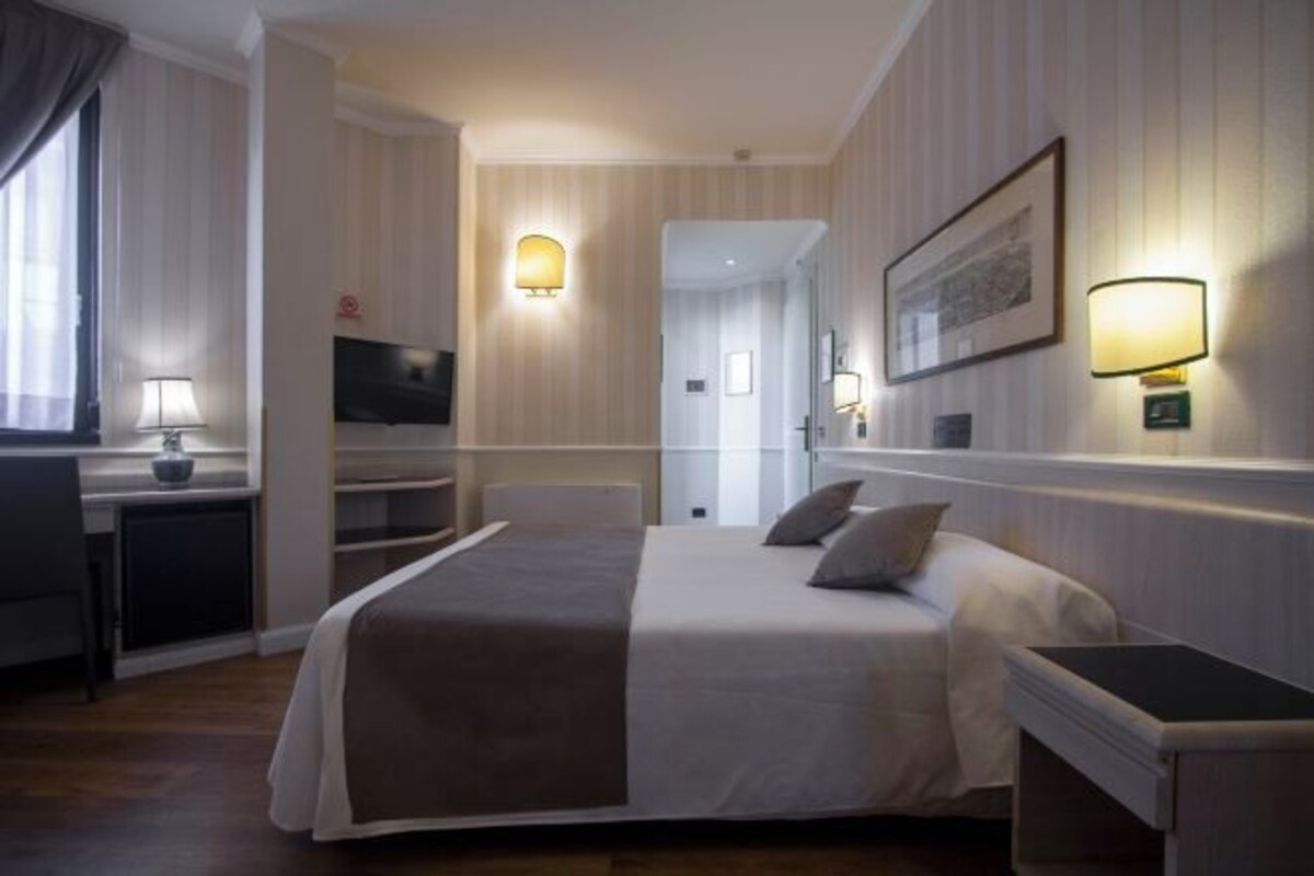 A hotel room with a bed, bedside tables, a desk, a TV, and wall-mounted lamps.