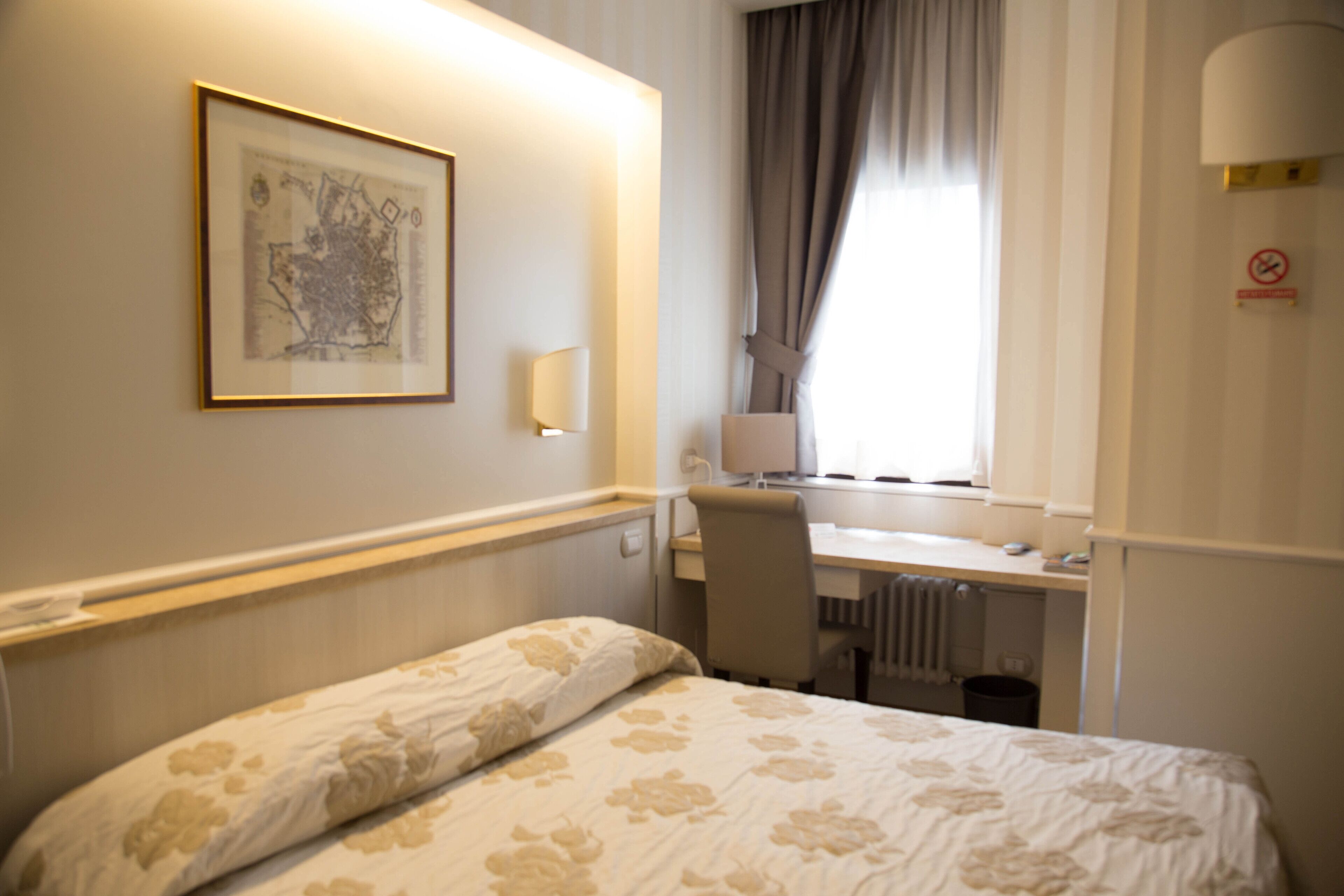 Ein Hotelzimmer mit Bett, Schreibtisch, Stuhl, Heizkörper, einem Fenster mit Vorhängen, einer Wandlampe und einer gerahmten Karte an der Wand.