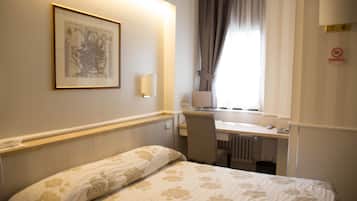 A hotel room with a bed, a desk, a chair, a radiator, a window with curtains, a wall-mounted lamp, and a framed map on the wall.