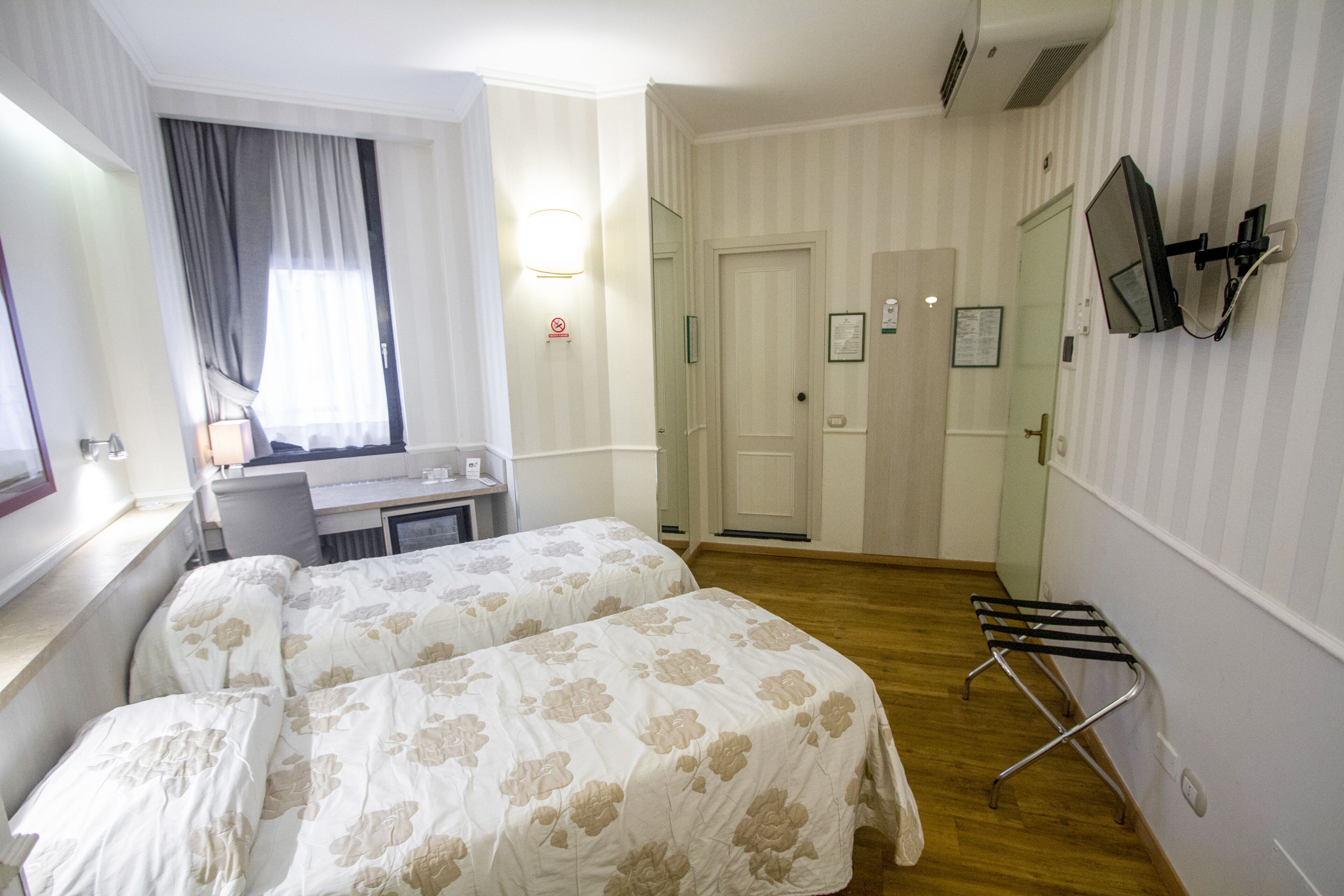 Camera d'albergo con due letti, una scrivania, una sedia, una TV a parete e una finestra con tende.