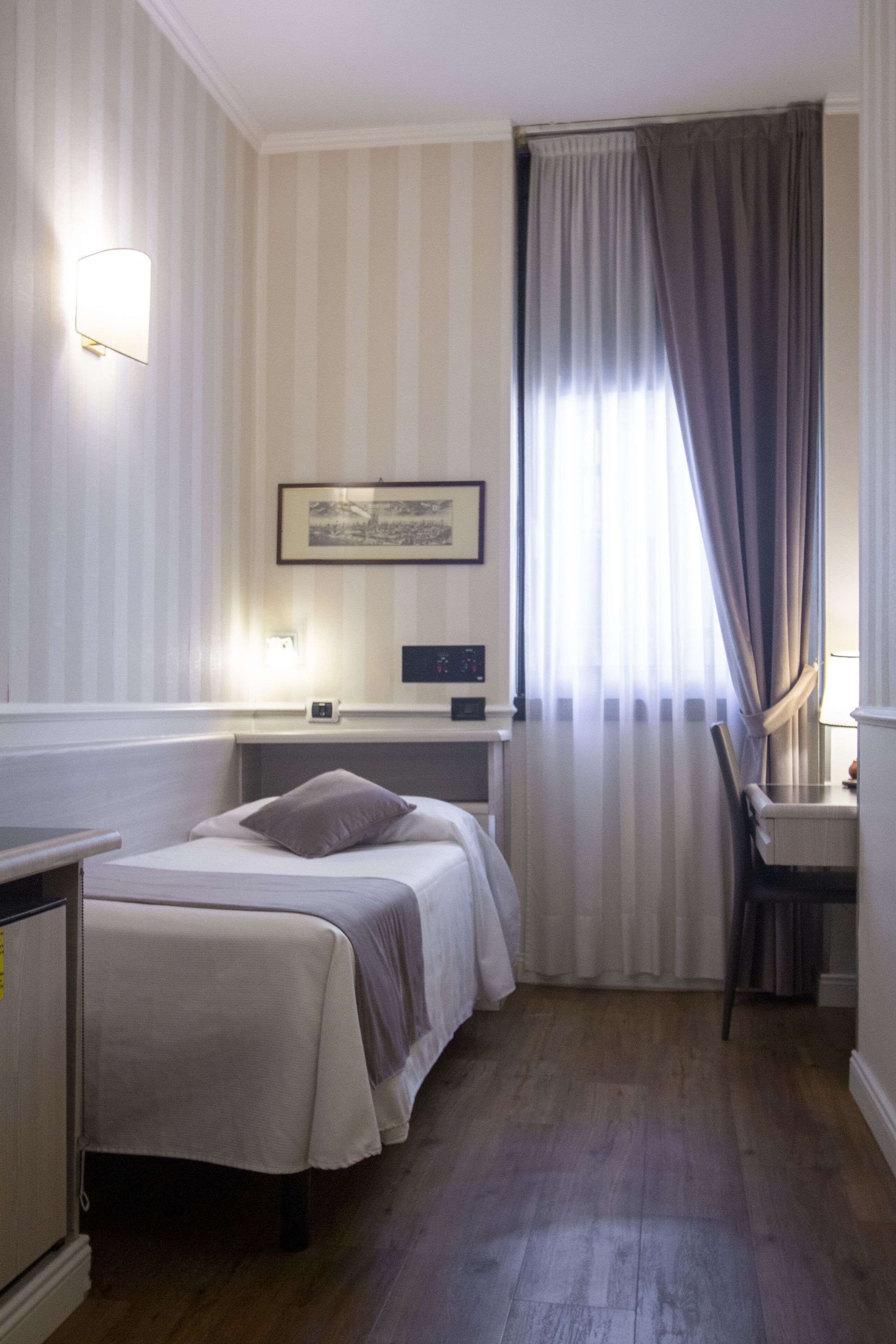 Una habitación de hotel con cama, mesita de noche, lámpara y ventana con cortinas.