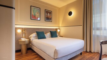 Camera d'albergo con un letto grande, due applique, un comodino rotondo con un telefono e una lampada, e due poster incorniciati sulla parete.