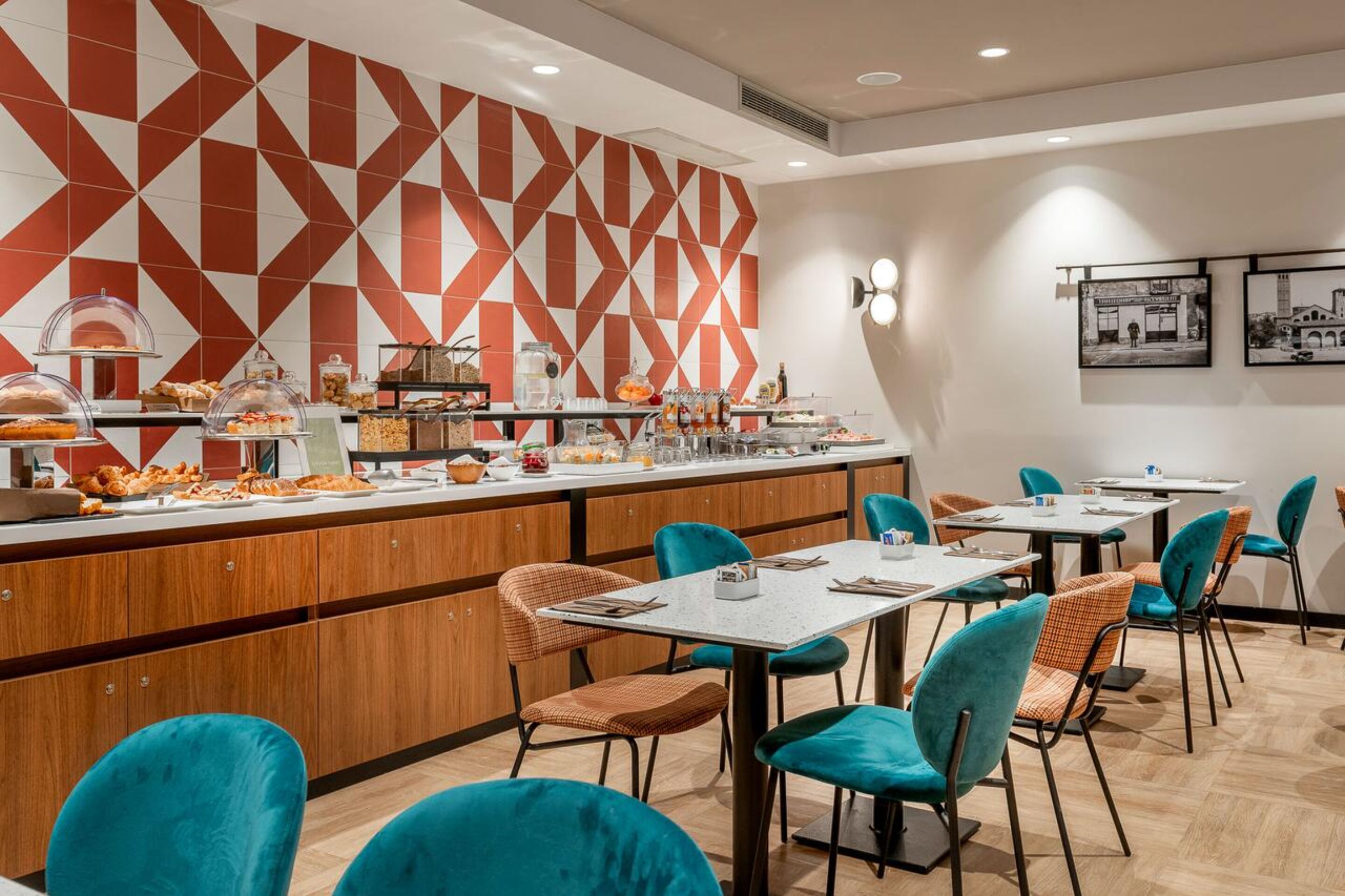 A modern restaurant with a geometric patterned wall, buffet counter with various food items, and teal chairs around tables.