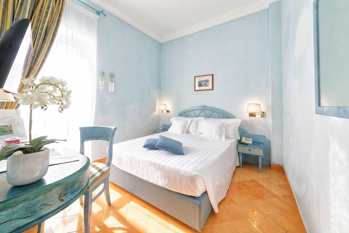 Habitación de hotel con cama, mesitas de noche, escritorio con silla, planta en maceta y ventana con cortinas.