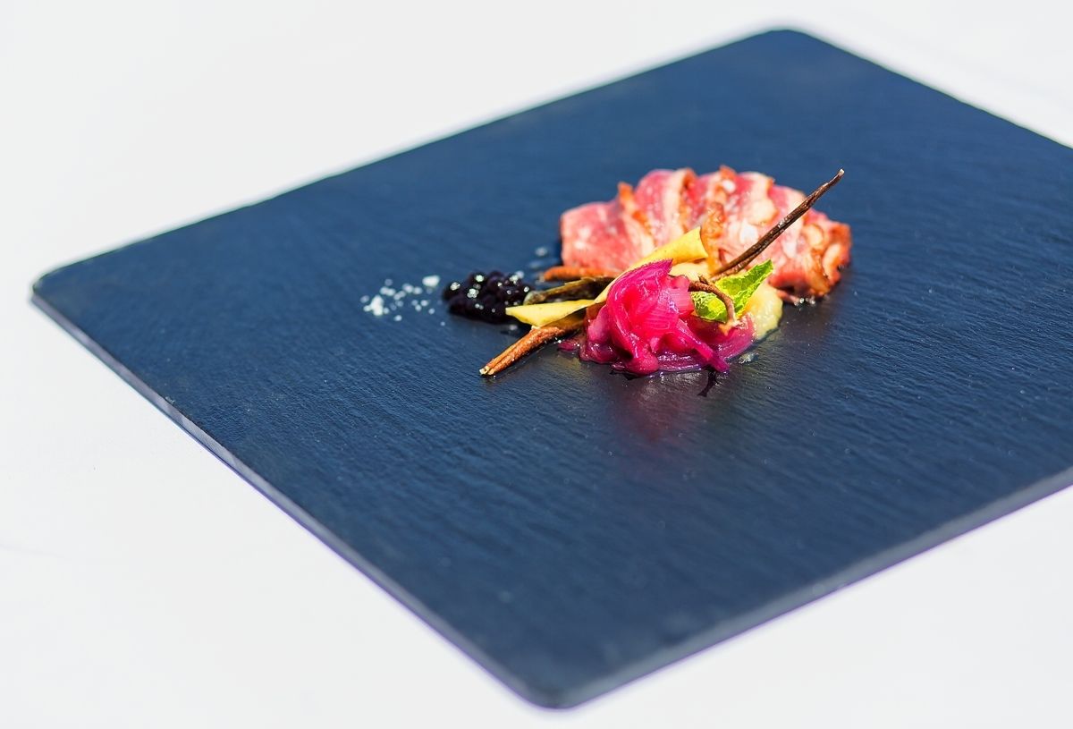 Une assiette carrée à fond bleu foncé, contenant un morceau de viande rose, quelques mûres et une garniture d’oignon rouge et d’herbes vertes.