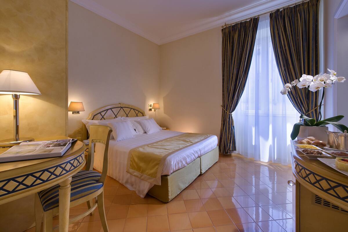 A hotel room with a bed, a desk with a lamp, a chair, and a small table with a vase of flowers.