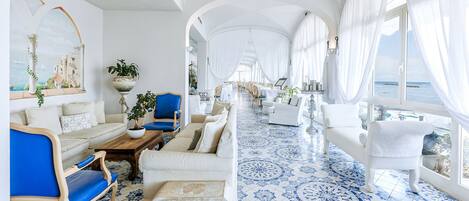 A spacious room with a blue and white tiled floor, white furniture, and large windows offering a view of the sea.