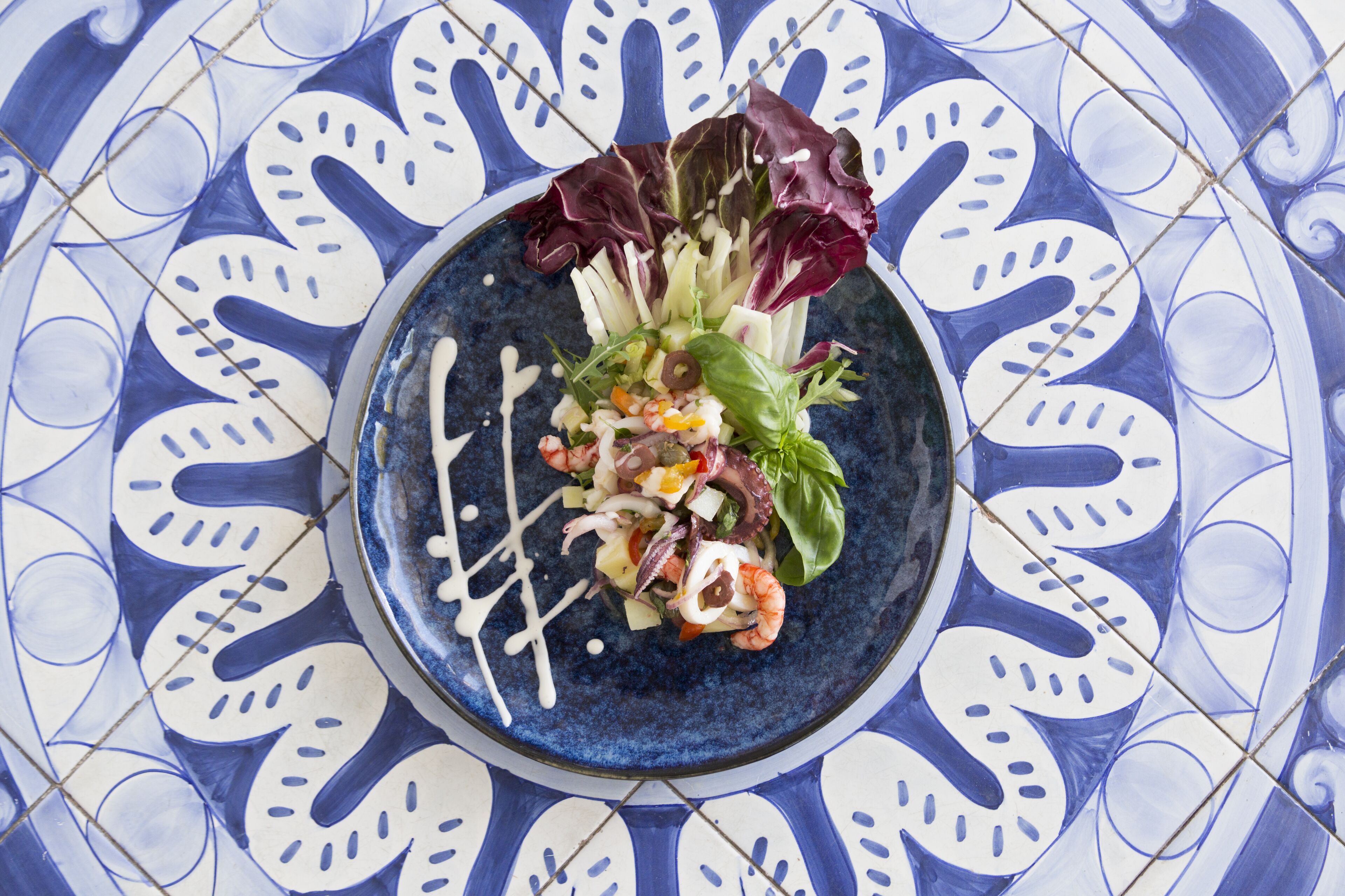 A plate of food with various vegetables and shrimp, served on a blue plate with a decorative tile background.