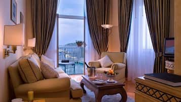 Habitación de hotel con sofá, sillón y una pequeña mesa con frutero y vasos. Un televisor está sobre un mueble de madera, y se ve un balcón con piscina a través de la ventana.