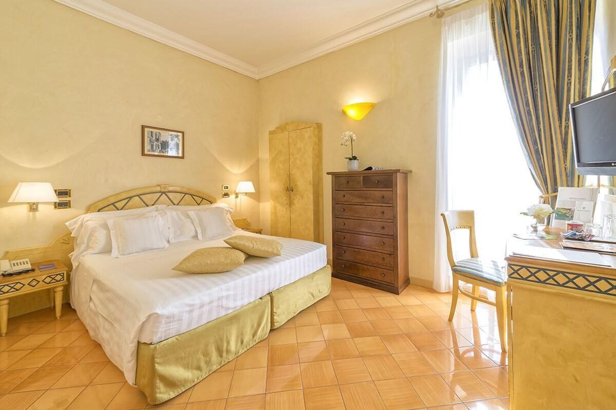 A hotel room with a bed, bedside lamps, a dresser, a desk with a chair, a television, and a window with curtains.