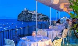 A seaside restaurant with tables set for dining, overlooking a rocky coastline with a historic building. - Miramare E Castello Hotel (Ischia)