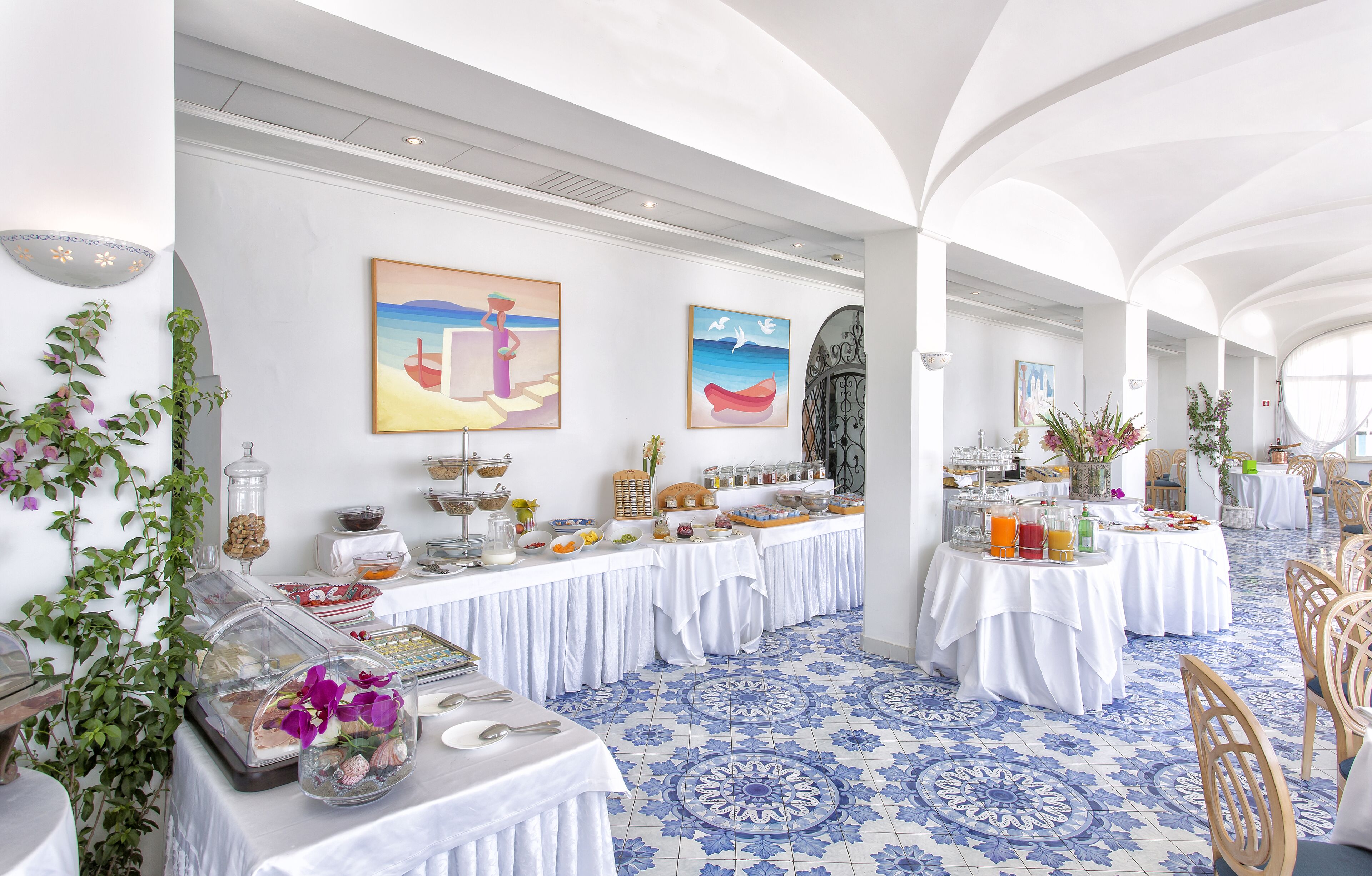 A spacious dining area with white tables, blue patterned floor tiles, and artwork on the walls.