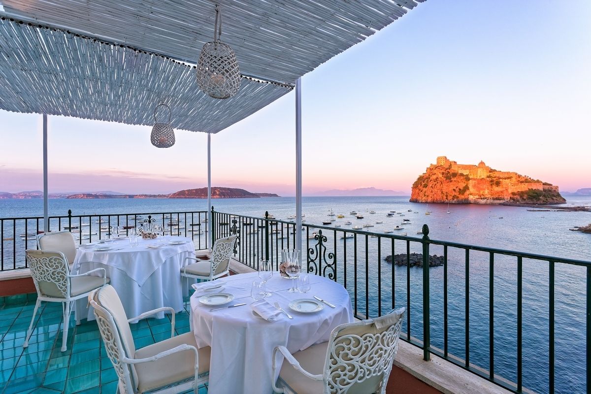 An outdoor dining area with tables set for two, overlooking a body of water with boats and a distant island.
