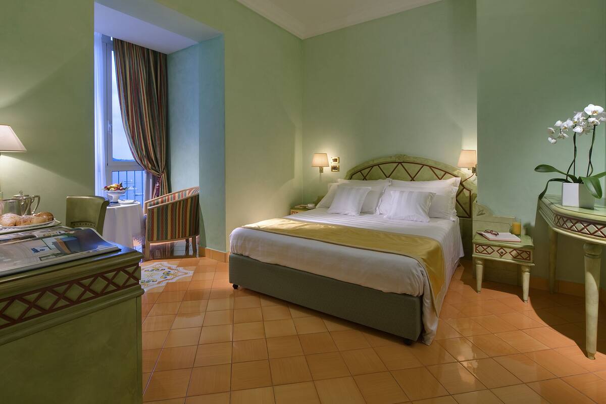 A hotel room with a bed, a desk with a lamp, a chair, a small table with a fruit bowl, and a window with curtains.