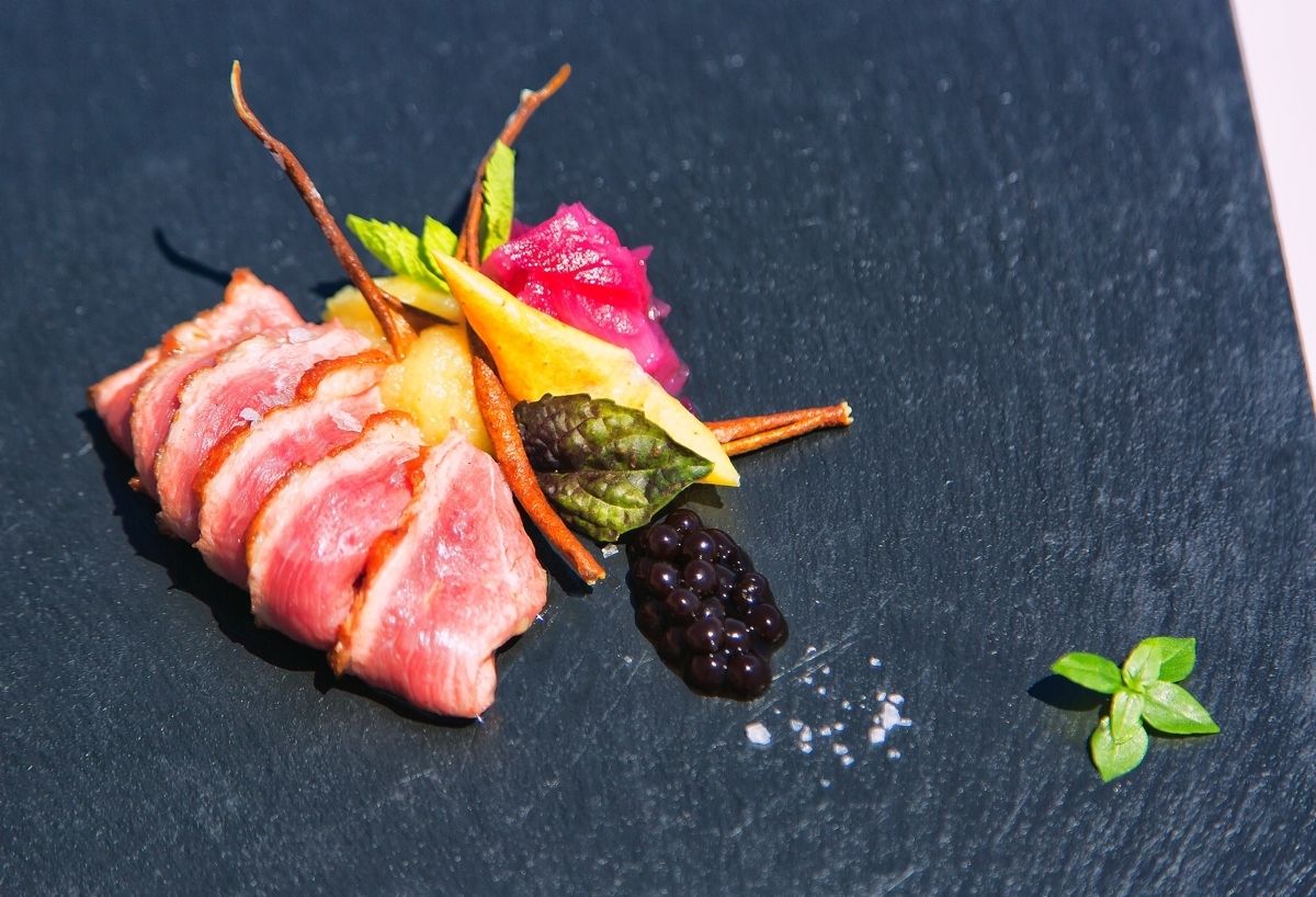 A dish with sliced meat, vegetables, and berries on a slate plate.