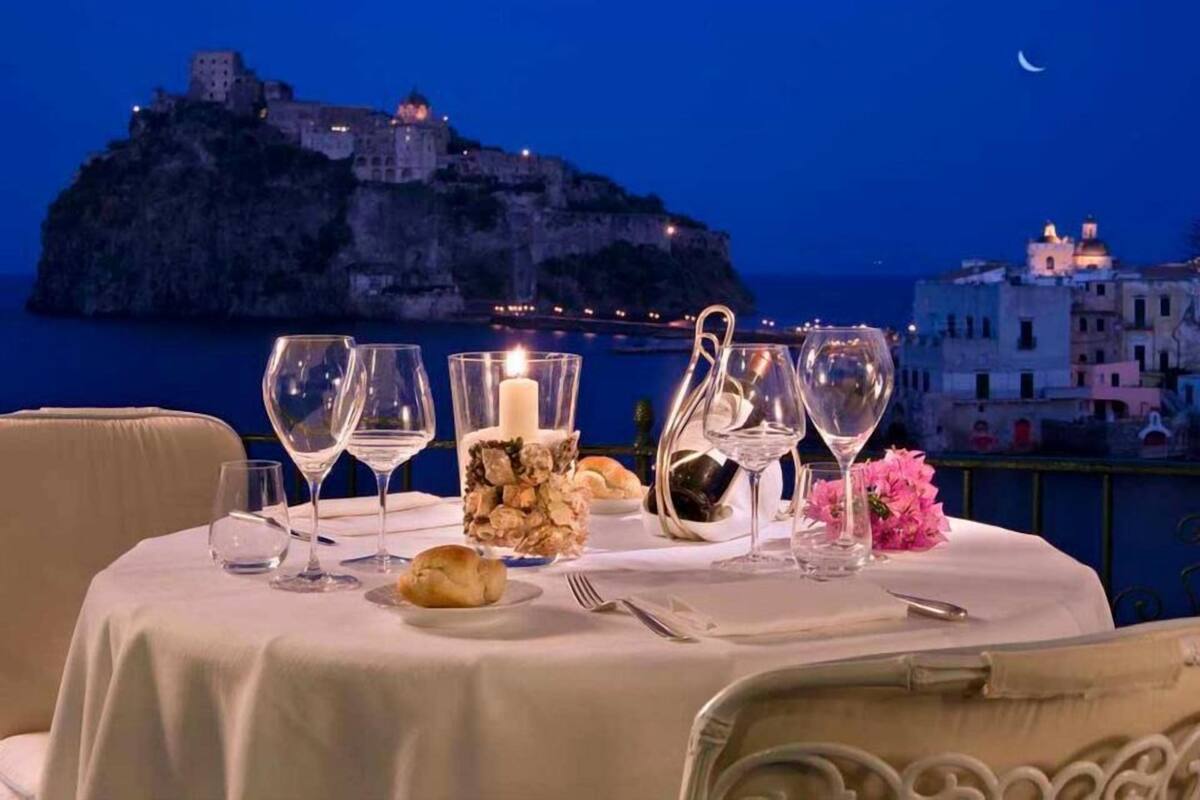 A table set for dining with a candle, wine glasses, and a bouquet of flowers. A scenic view of a coastal town with a castle on a cliff is visible in the background.