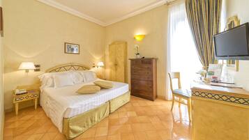 Une chambre d’hôtel comprenant un lit, des lampes de chevet, une commode en bois, un bureau avec une chaise et un téléviseur.