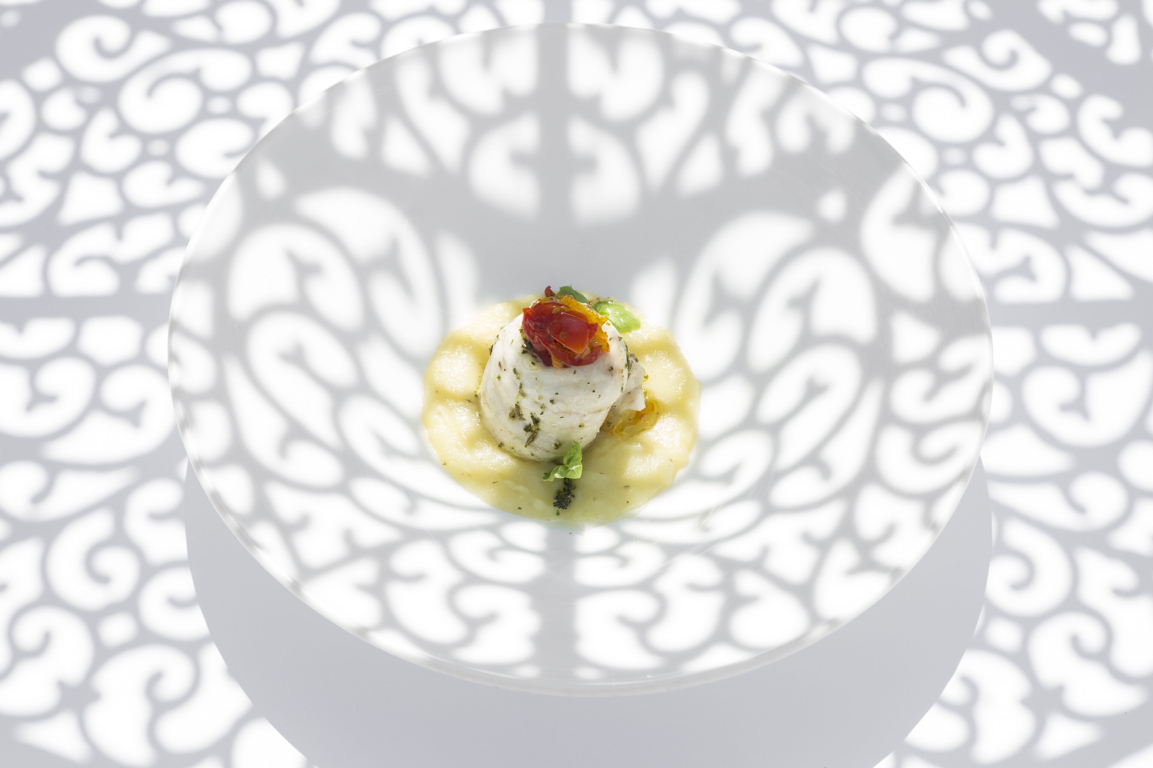 Une assiette blanche contenant un aliment blanc, peut-être du poisson, agrémenté d’herbes et surmonté d’une garniture rouge, le tout présenté sur un fond à motifs.