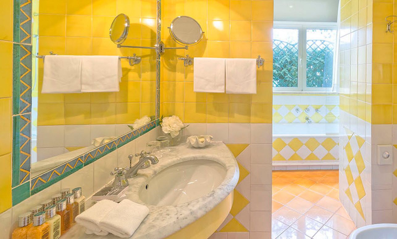 A bathroom with yellow tiles, a marble sink, two mirrors, and a window with a view of greenery.