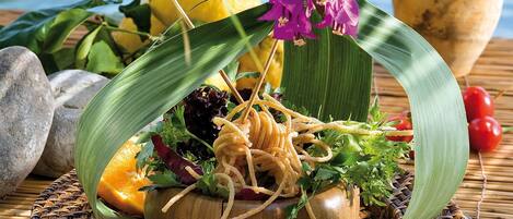 A wooden bowl filled with salad, garnished with purple flowers, placed on a woven mat.