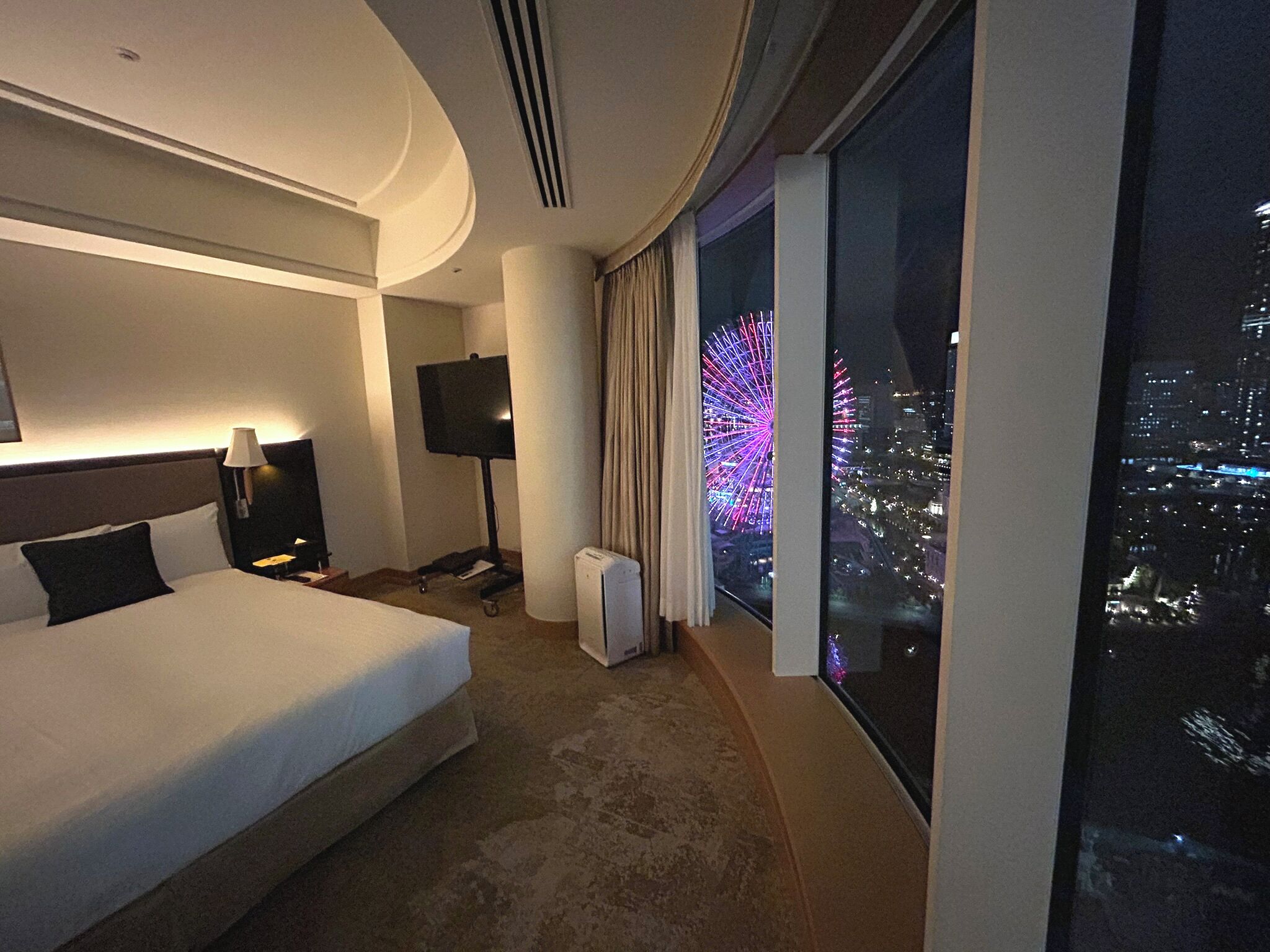 A hotel room with a large bed, a flat-screen TV, a heater, and a view of a cityscape with fireworks.