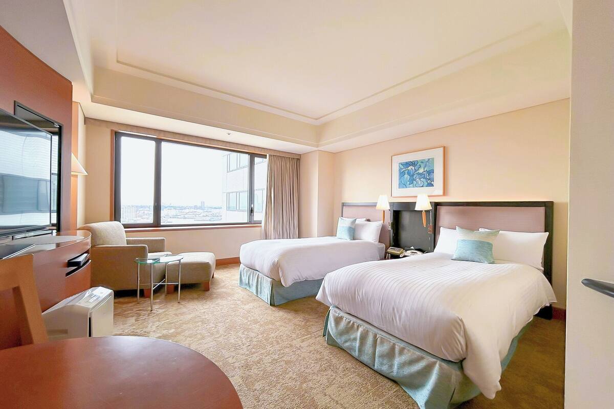 Habitación de hotel con dos camas, un escritorio, una silla, un televisor y un amplio ventanal con vista a la ciudad.