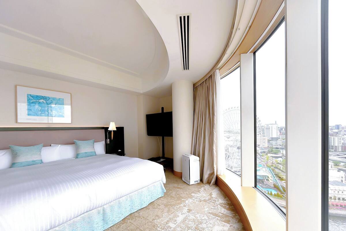 Habitación de hotel moderna con una cama grande, un televisor de pantalla plana, un respiradero en el techo y vistas a la ciudad a través de amplios ventanales.