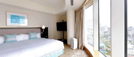 A modern hotel room with a large bed, a flat-screen TV, a ceiling vent, and a city view through large windows.