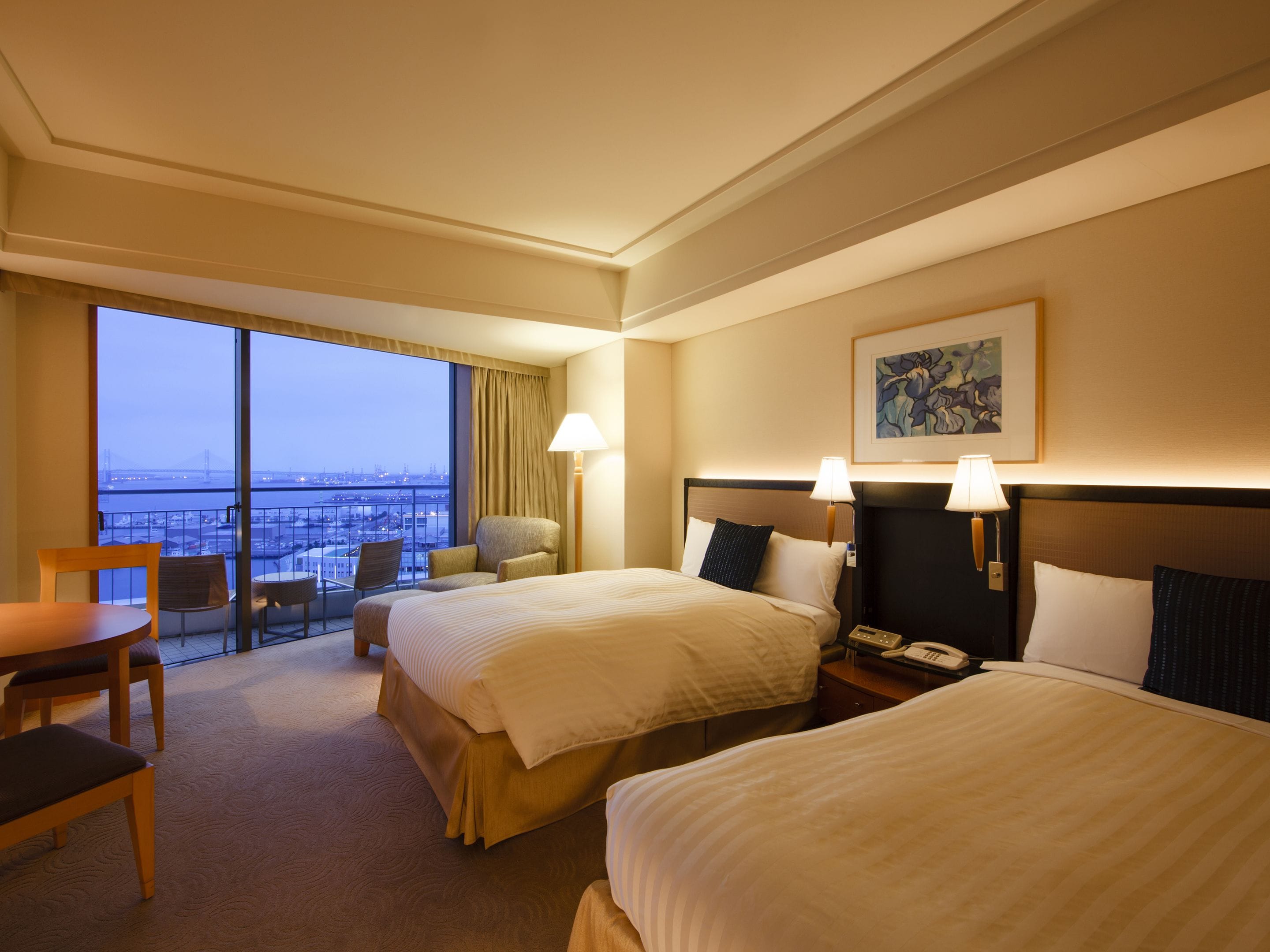 A hotel room with two beds, a balcony with a view of the city, a desk, and a chair.