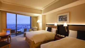 A hotel room with two beds, a balcony with a view of the city, a desk, and a chair.