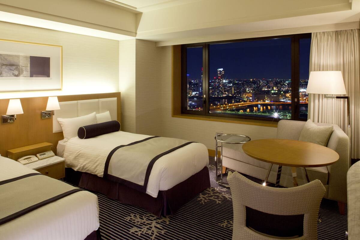 Habitación de hotel con dos camas, una mesita y vista a un paisaje urbano de noche.