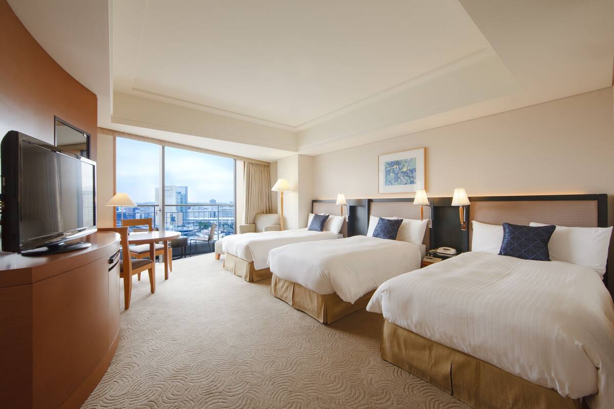 Habitación de hotel con dos camas, televisión, escritorio y ventana grande con vista a la ciudad.
