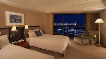 Ein Hotelzimmer mit zwei Betten, einem Schreibtisch, einem Stuhl und einem großen Fenster mit Blick auf die Stadt.