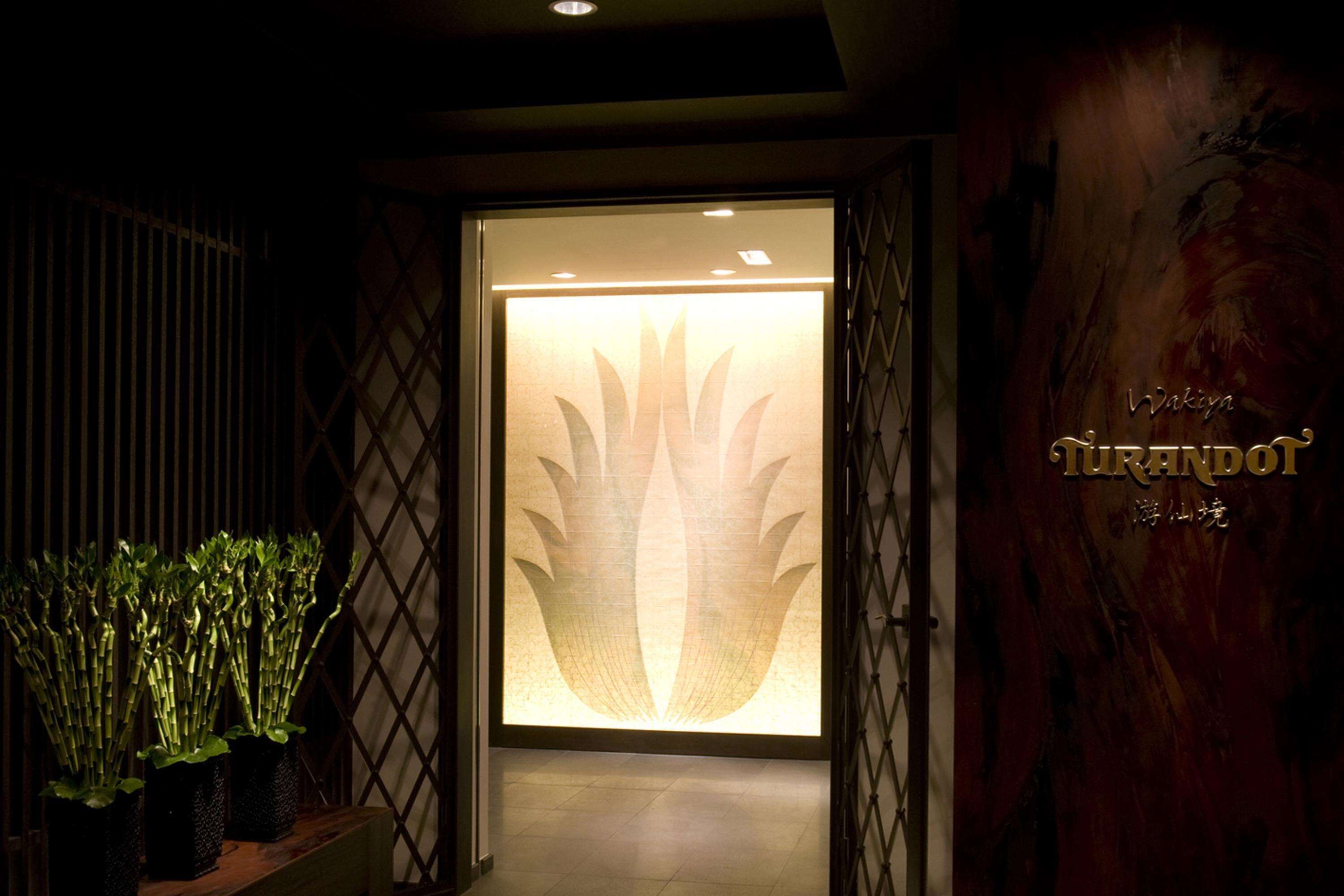 Un área de recepción con diseño moderno, que presenta un gran motivo de flor de loto iluminada en la puerta, un mostrador de recepción de madera y un cartel con el nombre 'Wakiyu'.