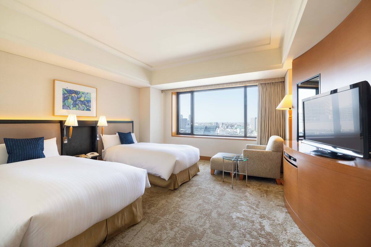 Habitación de hotel con dos camas, televisión, escritorio, silla y un amplio ventanal con vistas a la ciudad.