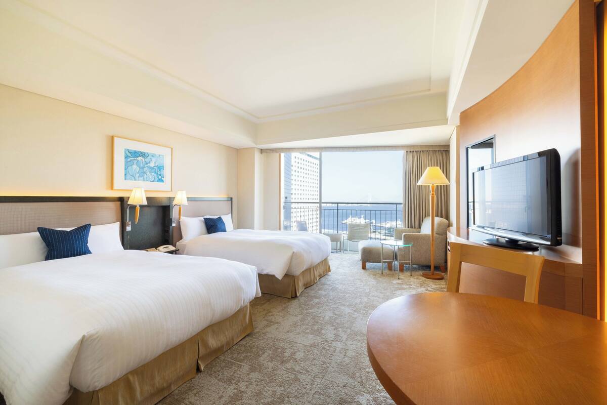 Habitación de hotel con dos camas, televisión, escritorio y balcón con vista a la ciudad.