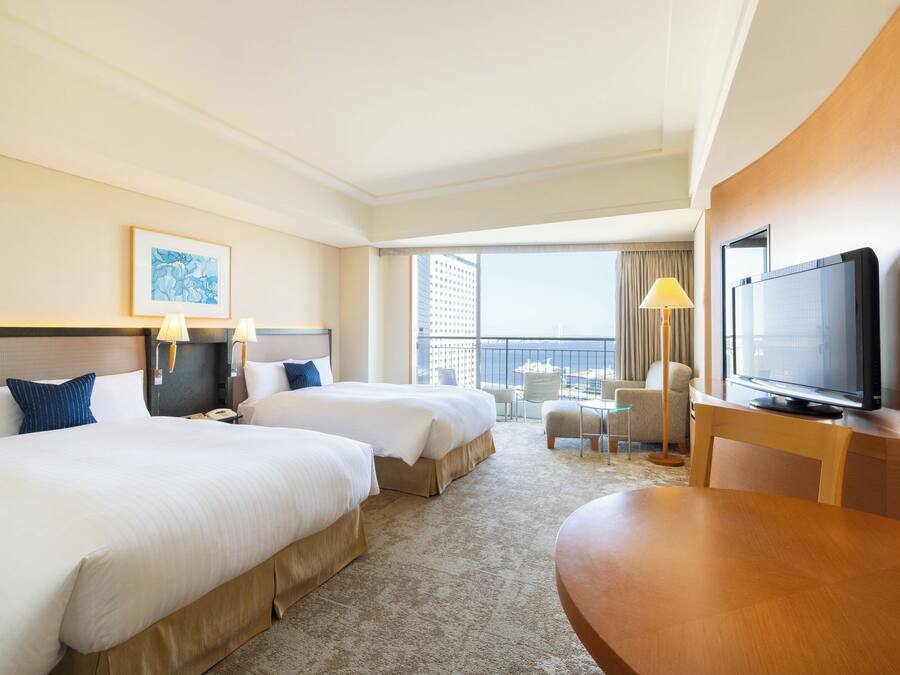 A hotel room with two beds, a television, a desk, and a balcony with a view of the city.