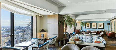 Amplia zona de estar con techos altos, ventanas grandes con vistas a la ciudad y diferentes opciones de asientos, incluyendo sofás y sillones.