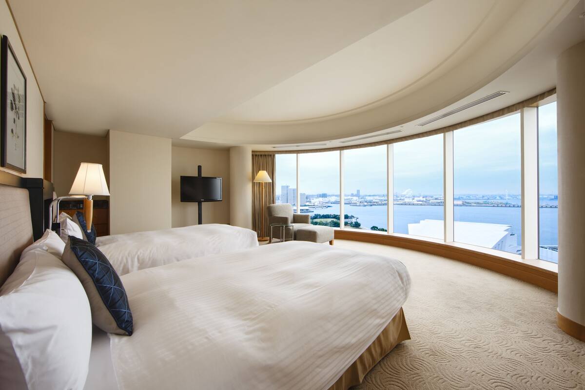 Habitación de hotel con dos camas, televisor, una silla y un ventanal con vista a la ciudad.