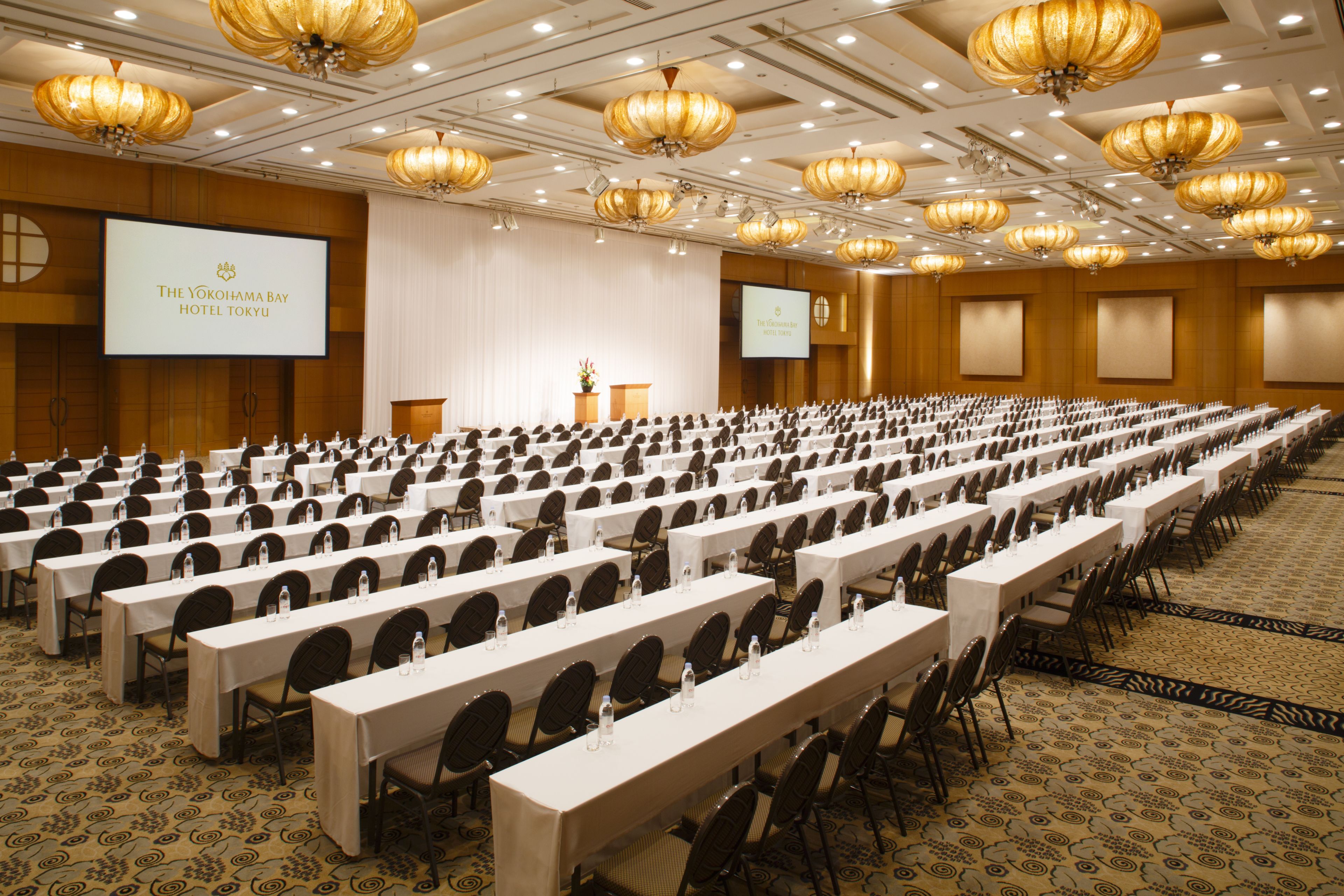 Un salón de conferencias en The Yokohama Bay Hotel Tokyo con filas de mesas y sillas, un atril y una pantalla grande.
