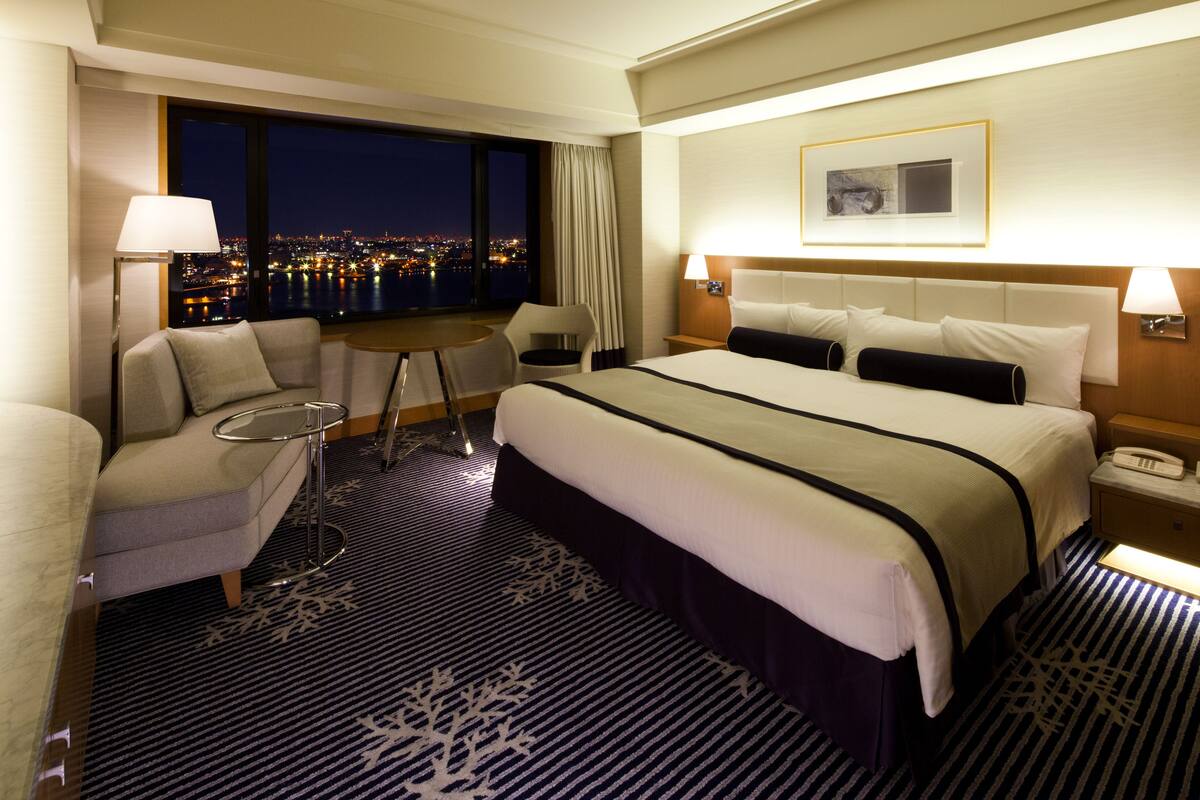 Habitación de hotel con una cama grande, un sofá, una mesita y vista a la ciudad de noche.