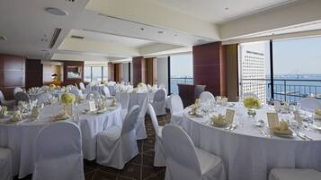 A banquet hall with round tables set for a formal event, large windows with a city view, and a high ceiling with recessed lighting.