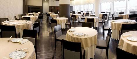 A spacious dining area with round tables, chairs, and a city view through large windows.