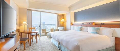 A hotel room with a large bed, a television, a desk with a chair, a small table, and a balcony with a view.
