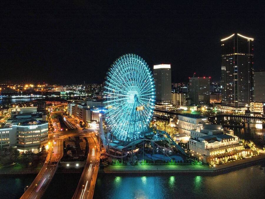 Una noria grande con estructura iluminada en azul, rodeada de edificios modernos y un muelle.